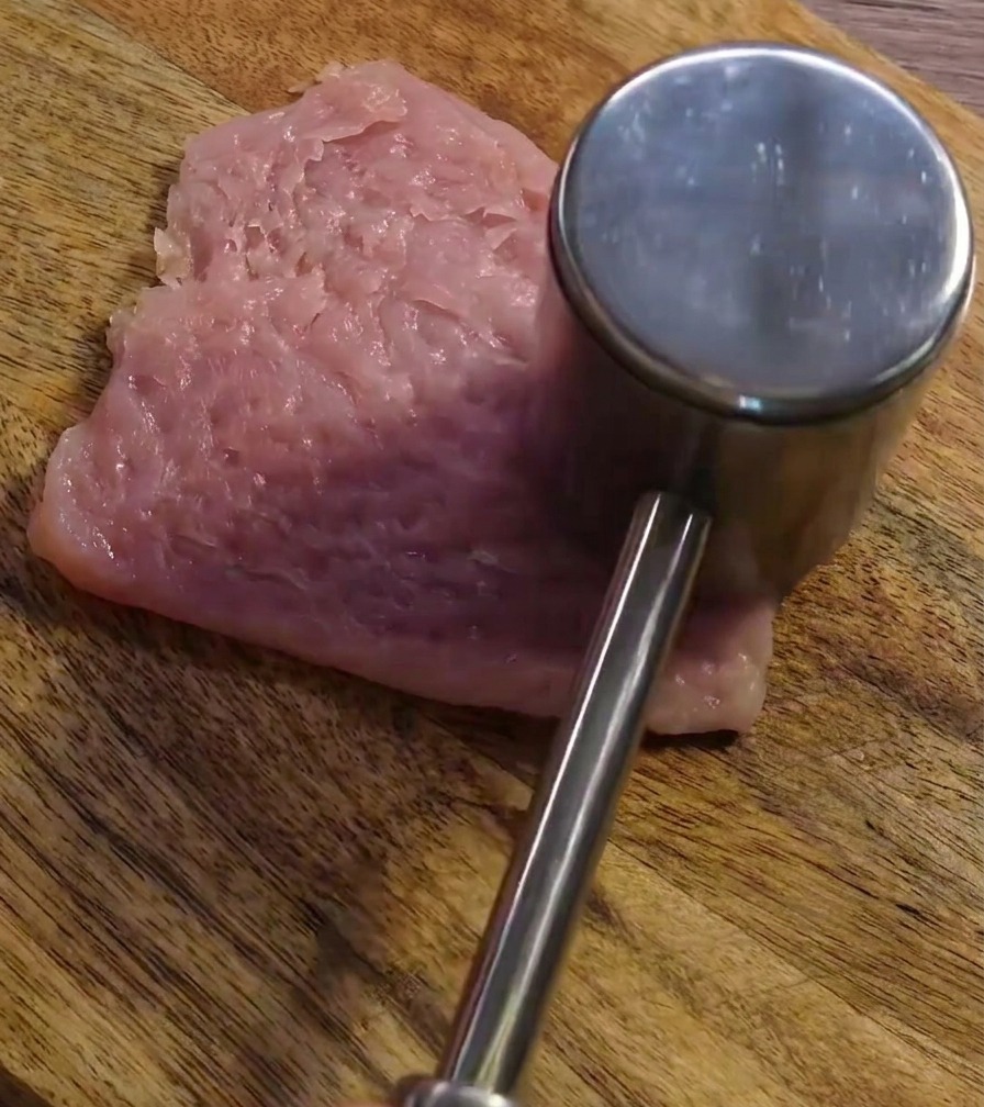 A metal meat mallet pounding a raw pork slice on a wooden board to flatten and tenderize it.