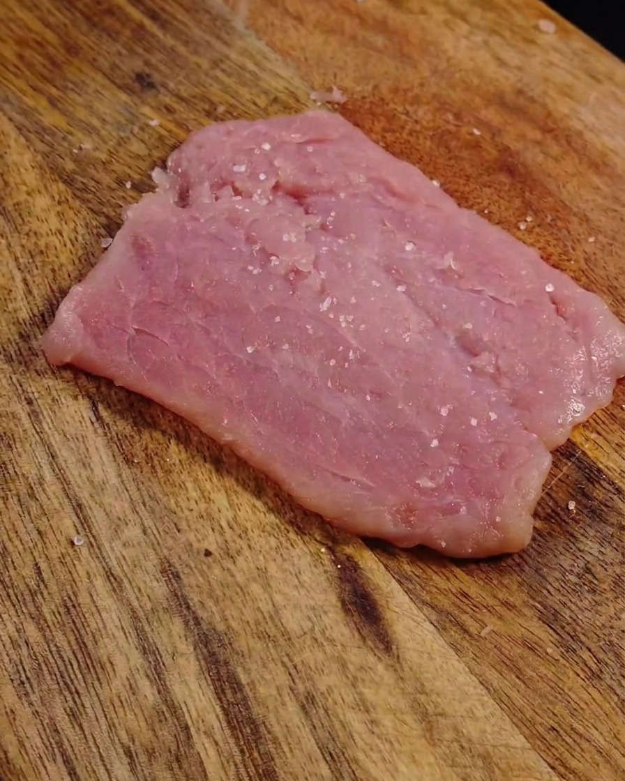 Salt and black pepper grains visible on the surface of a flattened, tenderized raw pork slice.