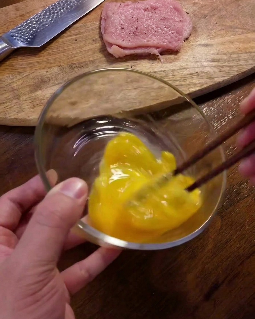 A person using dark brown chopsticks to whisk yellow raw eggs in a clear glass bowl on a wooden table.