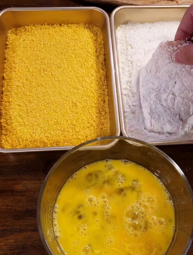A hand dredging a seasoned pork cutlet in a rectangular tray of white cornstarch powder.