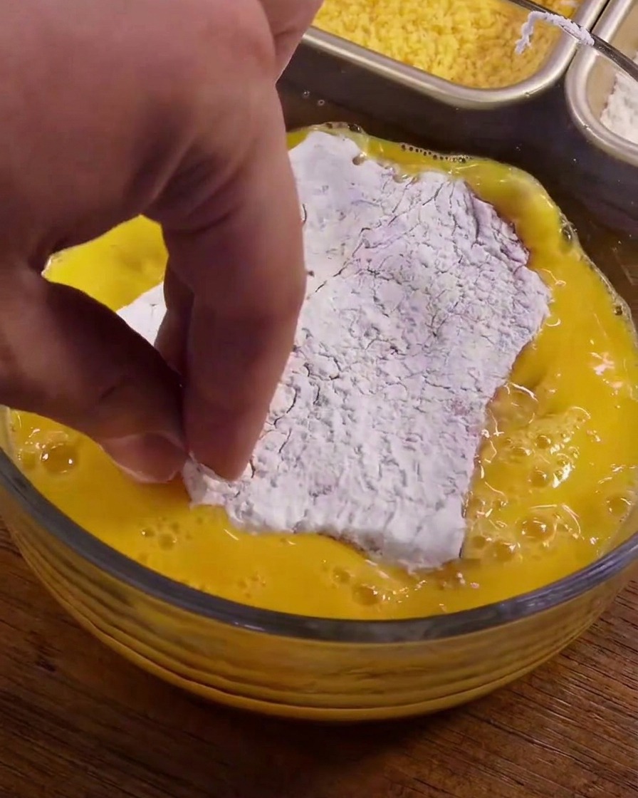 A piece of pork coated in white cornstarch being dipped into a bowl of yellow beaten eggs.