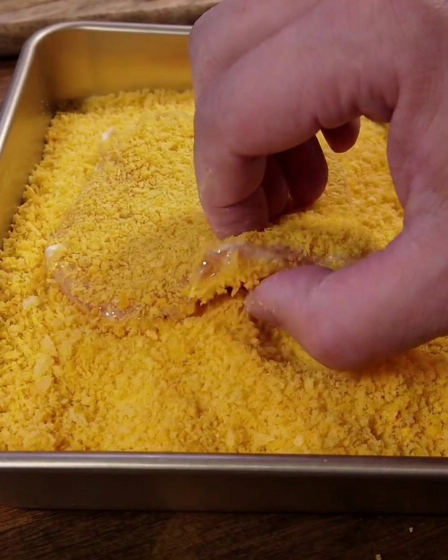 Pressing a piece of pork into a tray filled with golden-yellow Panko breadcrumbs.