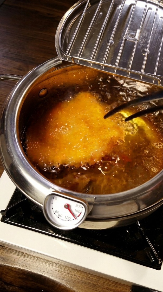 Breaded pork cutlet deep-frying in a pot of hot oil with a temperature probe visible.