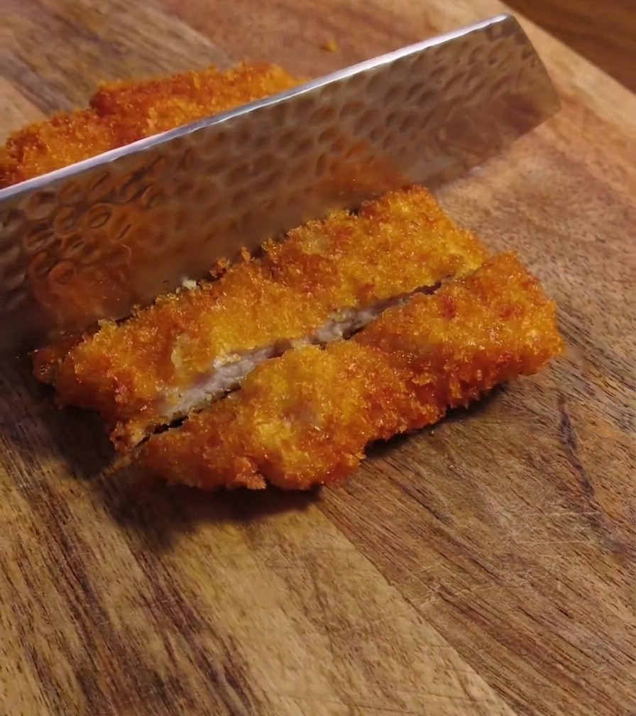 A close-up shot of a dimpled metal knife blade slicing through a crunchy, deep-fried pork cutlet on a wooden board.