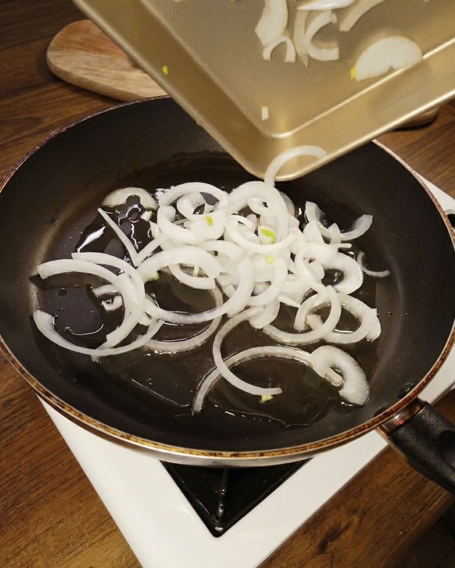 Sliced white onion rings being poured from a gold colored tray into a black non stick frying pan with shimmering oil