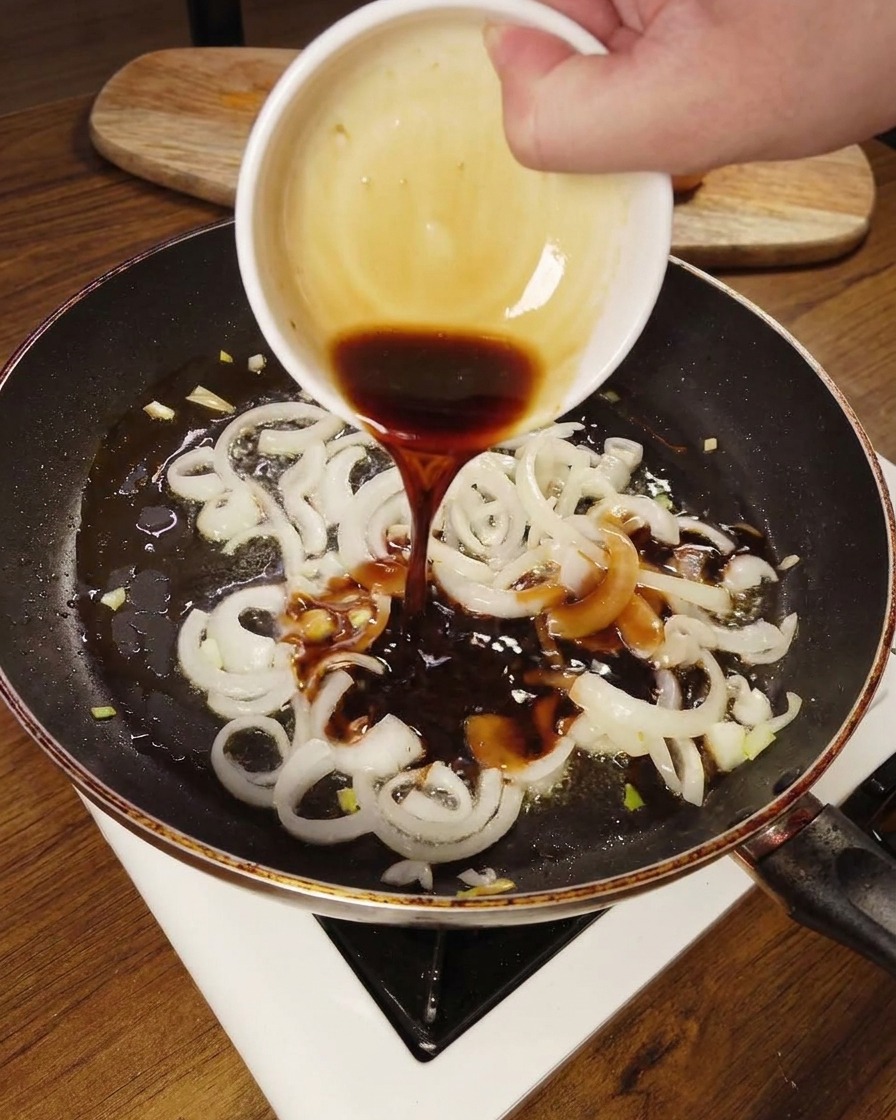Dark savory sauce being poured from a small white bowl onto softened white onions in a frying pan.