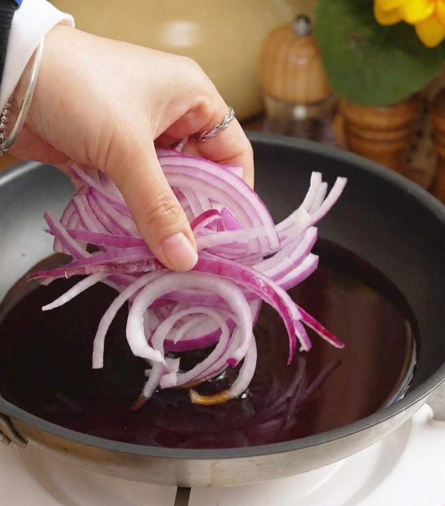 A hand dropping a heap of sliced red onions into a pan filled with simmering dark sauce.