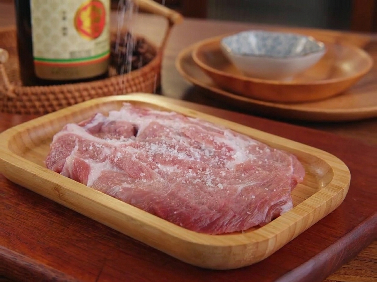 Fine salt and black pepper being sprinkled onto a raw piece of pork shoulder resting on a wooden tray.