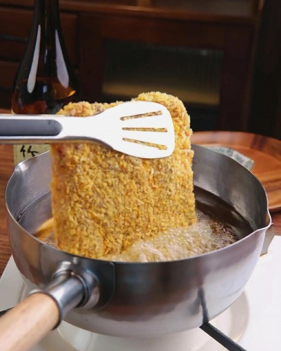 A breaded pork cutlet being lowered into a stainless steel pot of hot, bubbling frying oil using metal tongs.