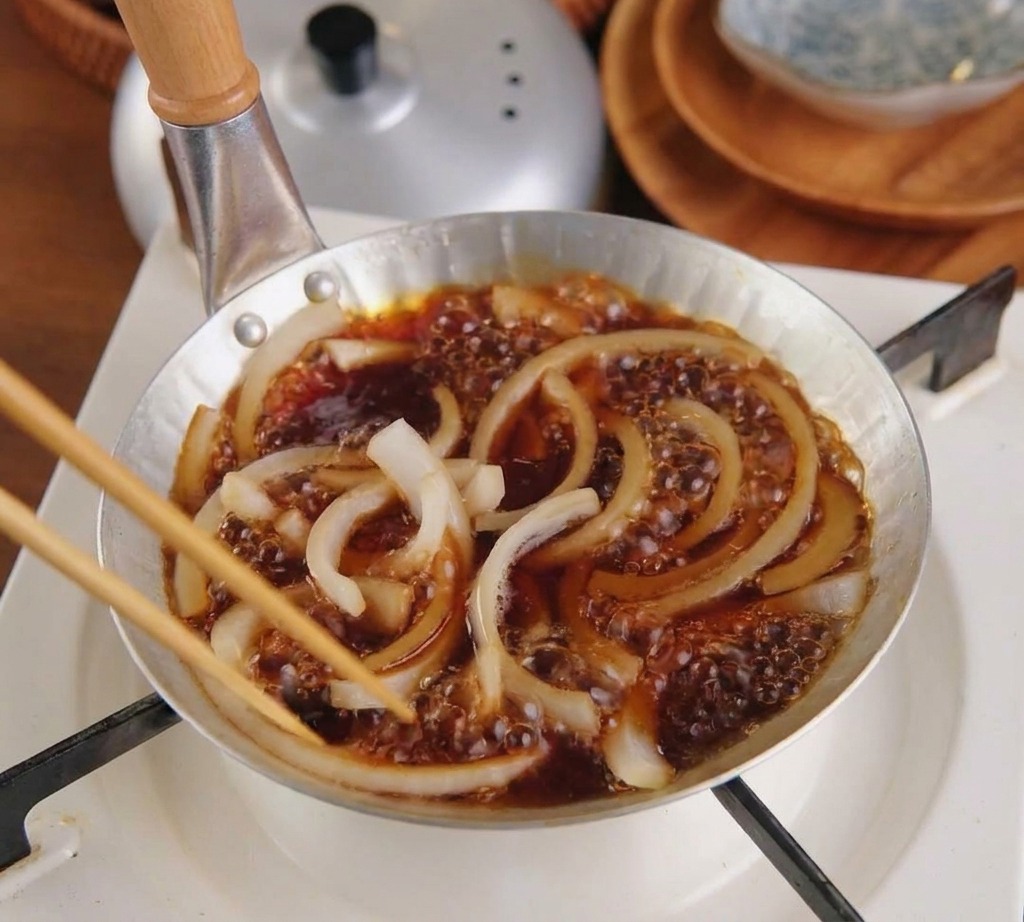 Thinly sliced white onions bubbling and simmering in a dark brown sauce inside a small silver pan on a portable gas stove.