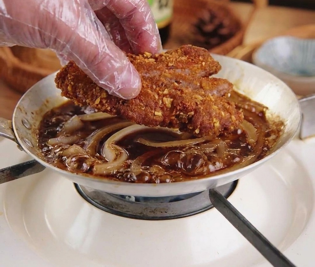 A gloved hand placing a sliced, crispy fried pork cutlet into a pan of simmering onions and dark seasoning sauce.