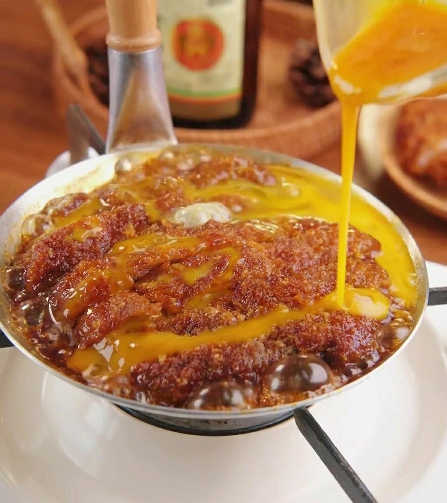 Whisked egg liquid being drizzled from a plastic bowl onto a pork cutlet simmering in a silver pan.