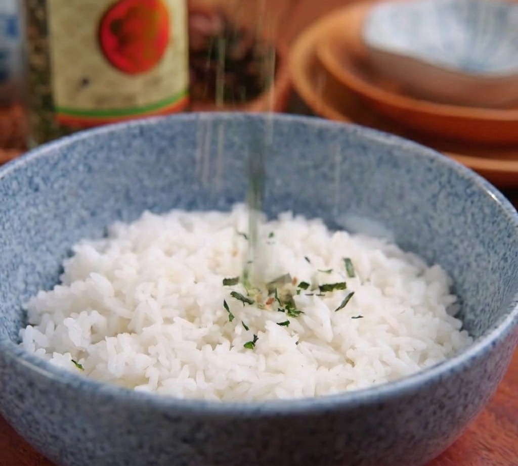 Dark green shredded nori seaweed flakes being sprinkled onto the center of a blue bowl filled with plain white rice.