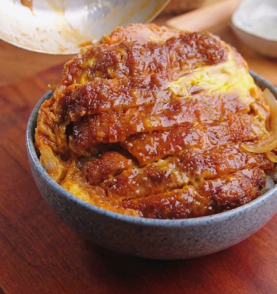 A pan being tilted over a rice bowl to slide a golden breaded pork cutlet topped with cooked eggs and onions onto the rice.