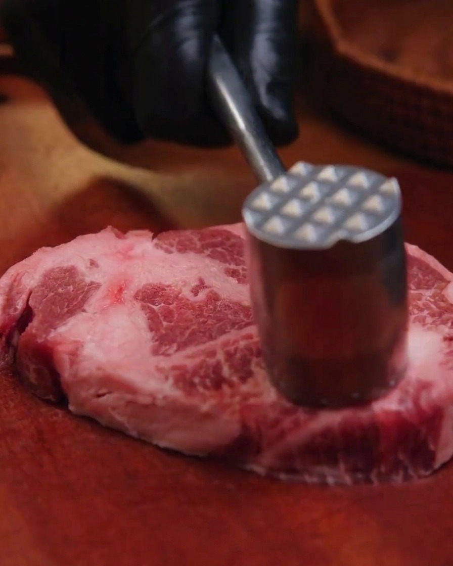 A hand in a black glove using a metal meat mallet to tenderize a thick slab of raw pork collar on a wooden board.