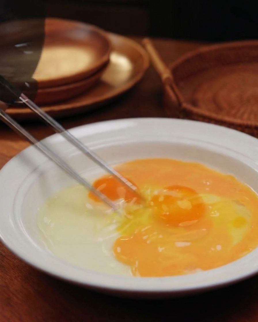 Chopsticks beating two eggs in a white rimmed bowl, creating a streaky yellow and orange mixture.