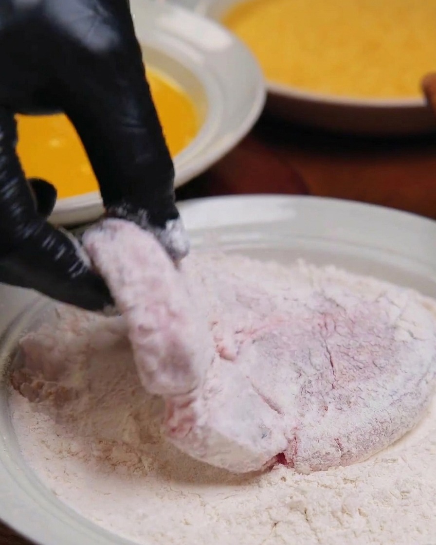 A gloved hand holding a thick piece of pork that has been completely coated in white flour over a shallow dish.