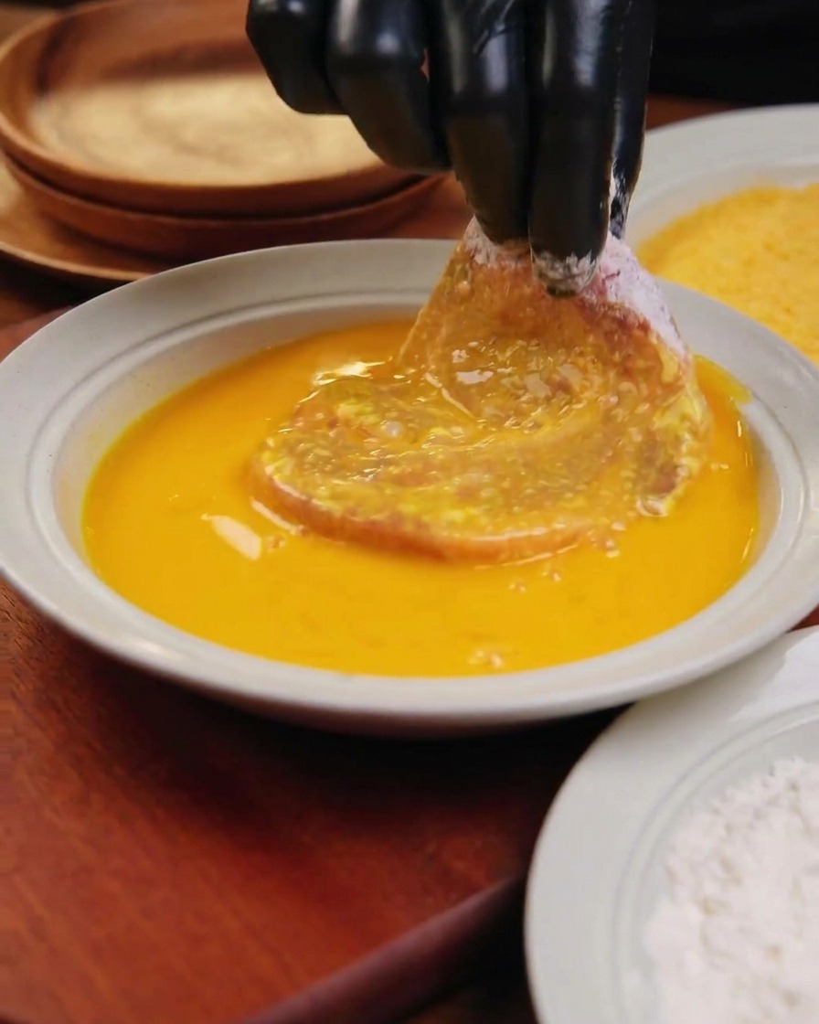 A hand in a black glove dipping a large, floured pork cutlet into a shallow white bowl filled with vibrant yellow egg wash.
