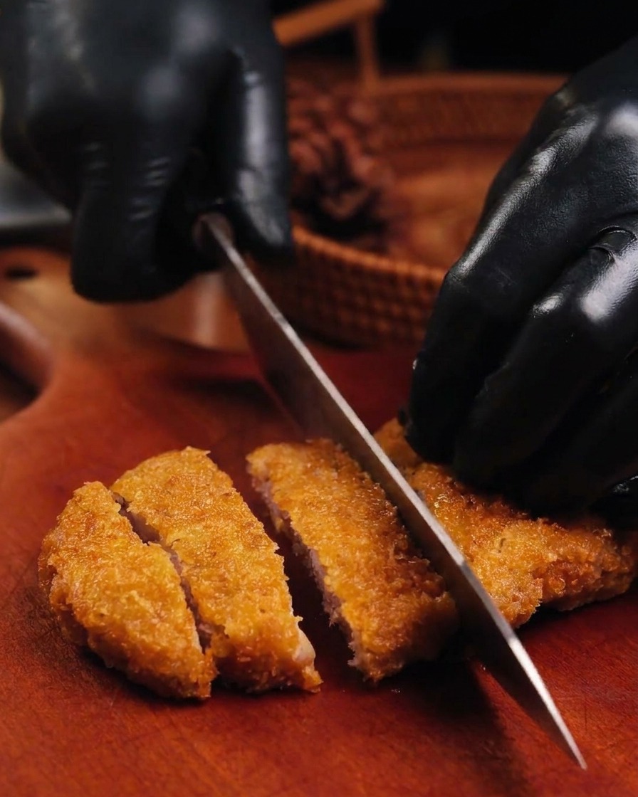 A sharp knife slicing through a crispy golden brown fried pork cutlet on a wooden board held by hands in black gloves.