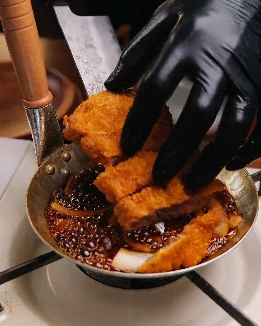 Hands in black gloves arranging sliced fried pork pieces into a small pan on top of a bed of simmering white onions and dark sauce.