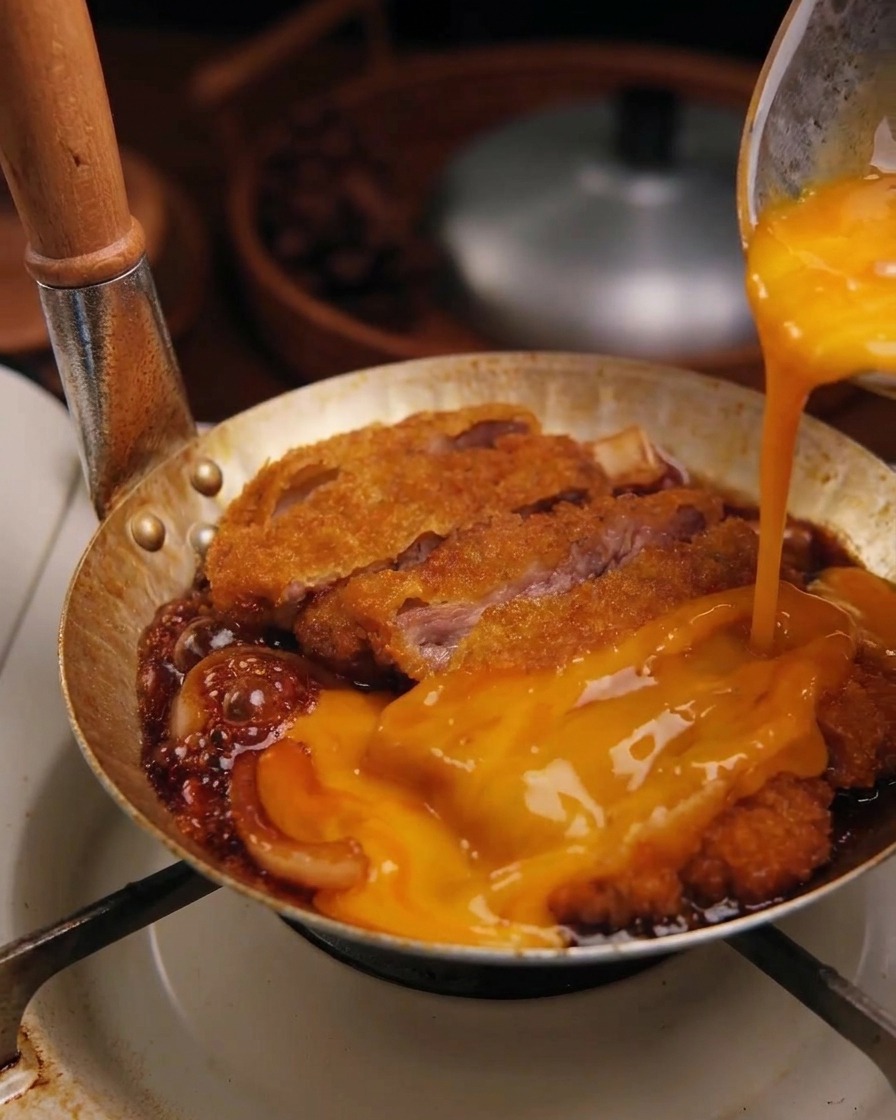 Beaten raw egg mixture being poured from a bowl onto a fried pork cutlet simmering in sauce with onions.