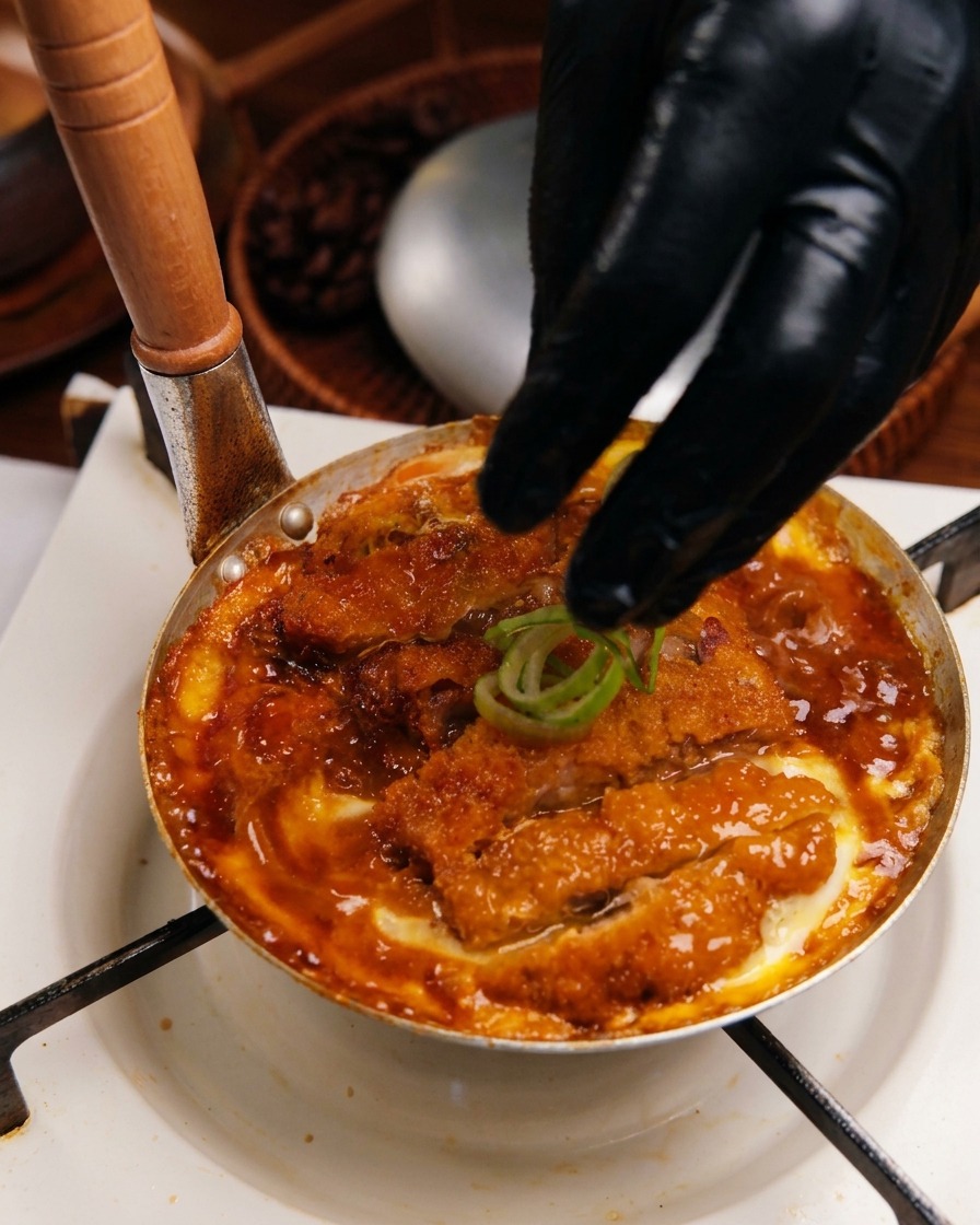 A gloved hand placing sliced green onion rings onto a cooked Katsudon mixture in a small metal pan.