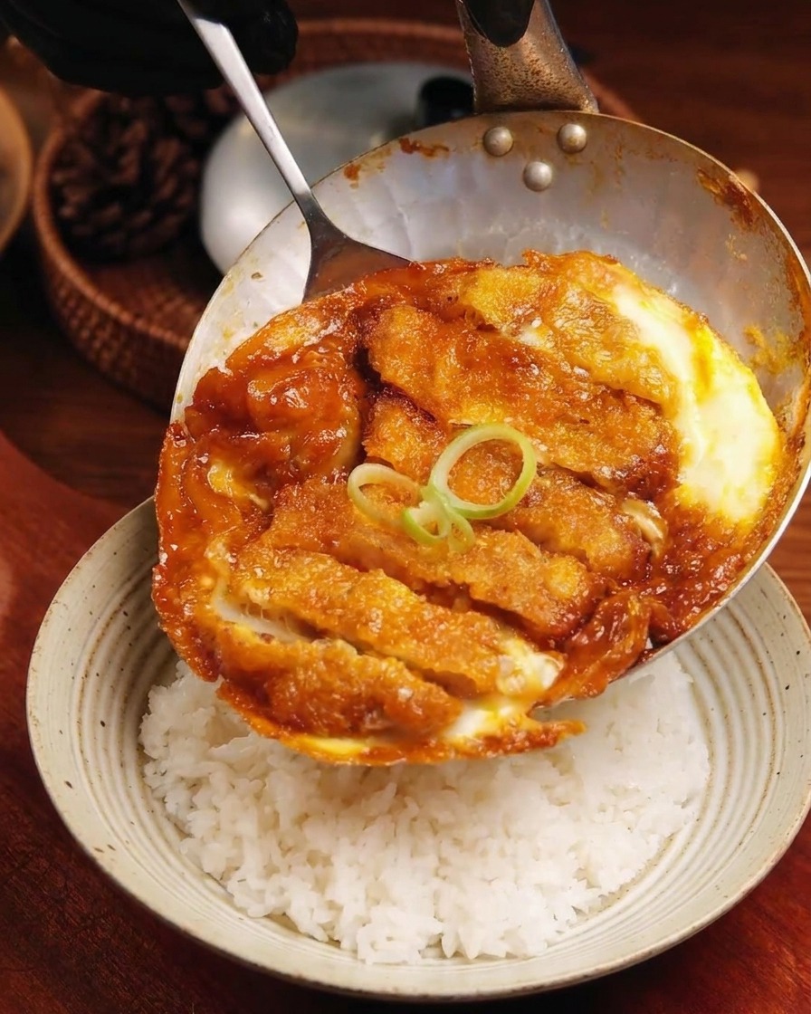 The completed pork cutlet and egg mixture being slid from a shallow pan onto a bowl of fluffy white rice.