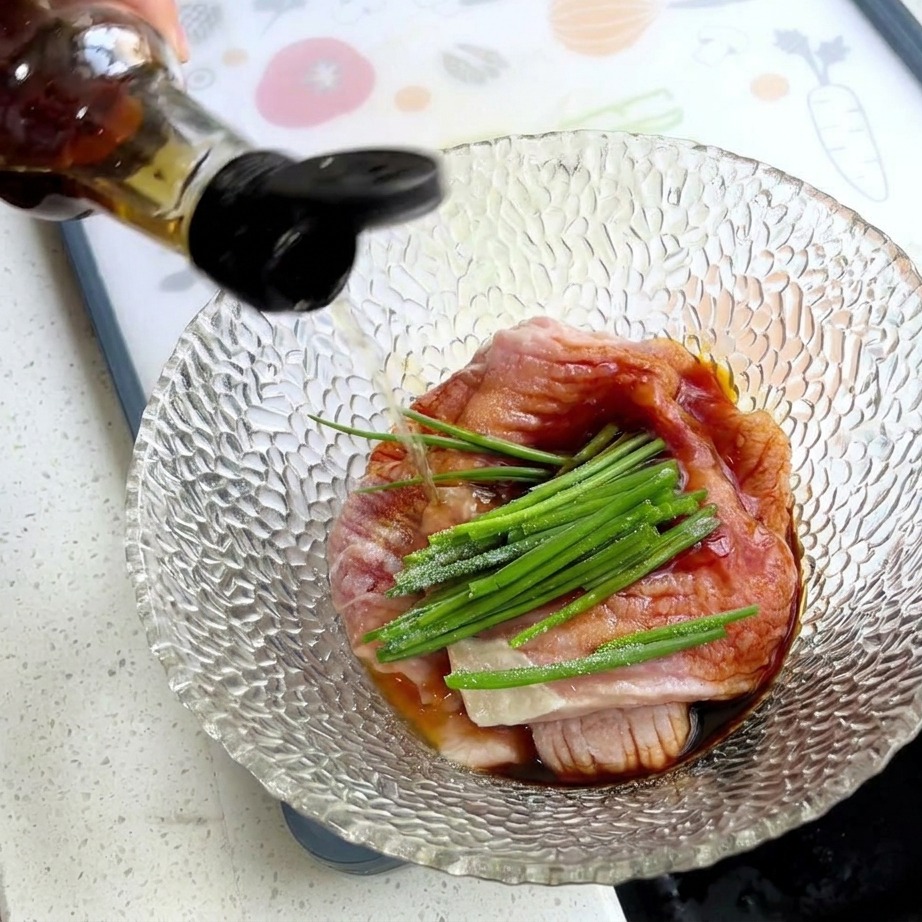 A hand pouring seasoning from a bottle into a bowl containing raw pork slices, green scallions, and white starch.