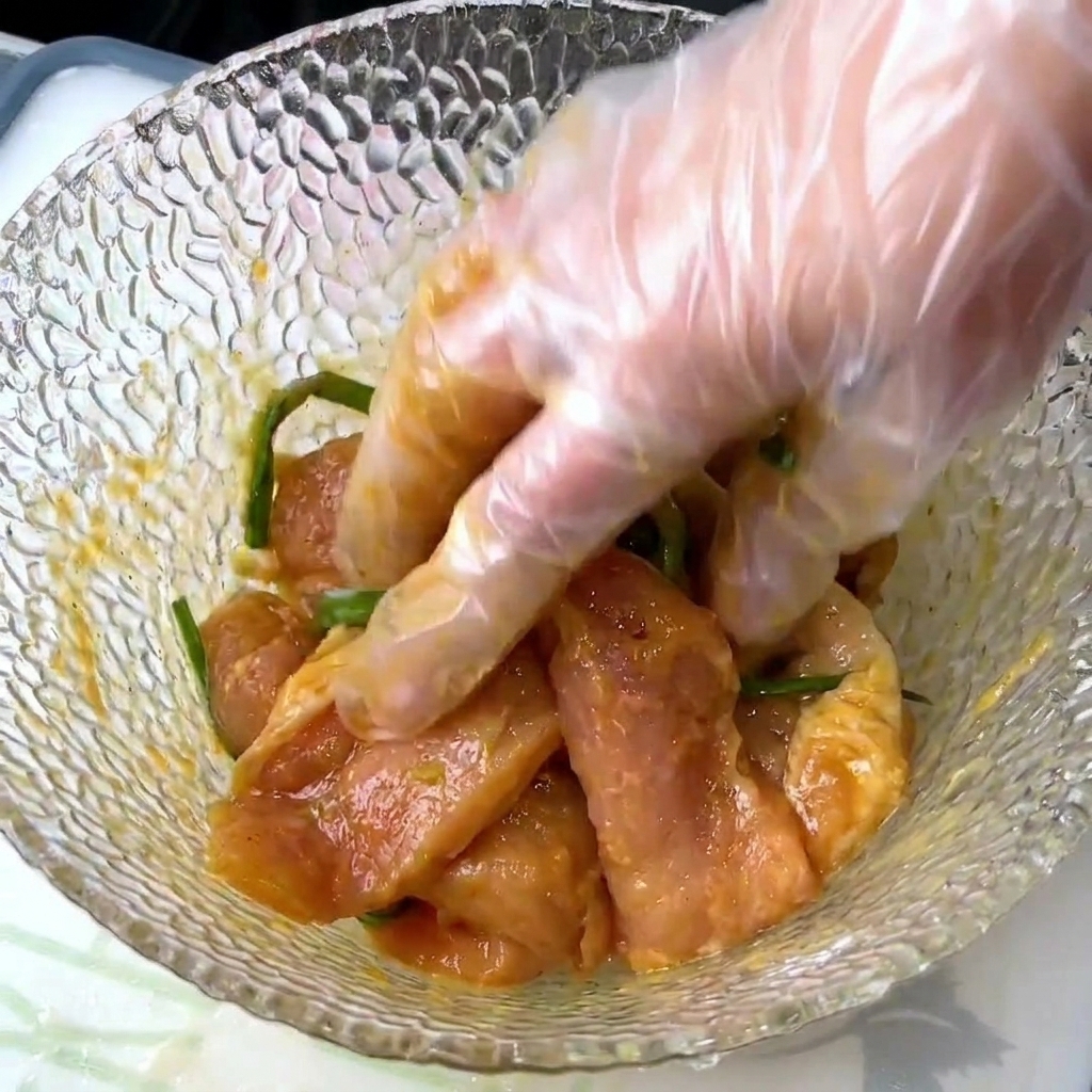 A gloved hand mixing raw pork slices with scallions and liquid seasonings in a textured glass bowl.
