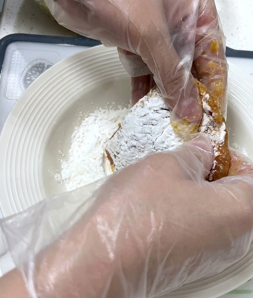 Gloved hands pressing a marinated pork cutlet into a shallow white plate filled with white starch powder.