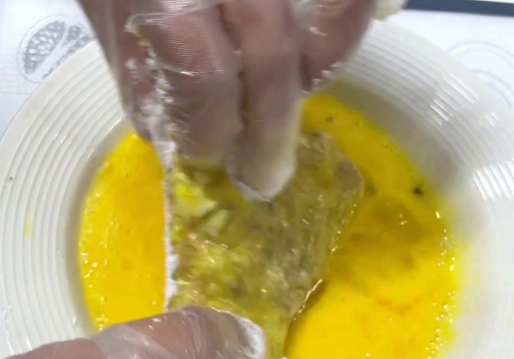Hands dipping a starch-coated pork cutlet into a plate of vibrant yellow beaten egg wash.