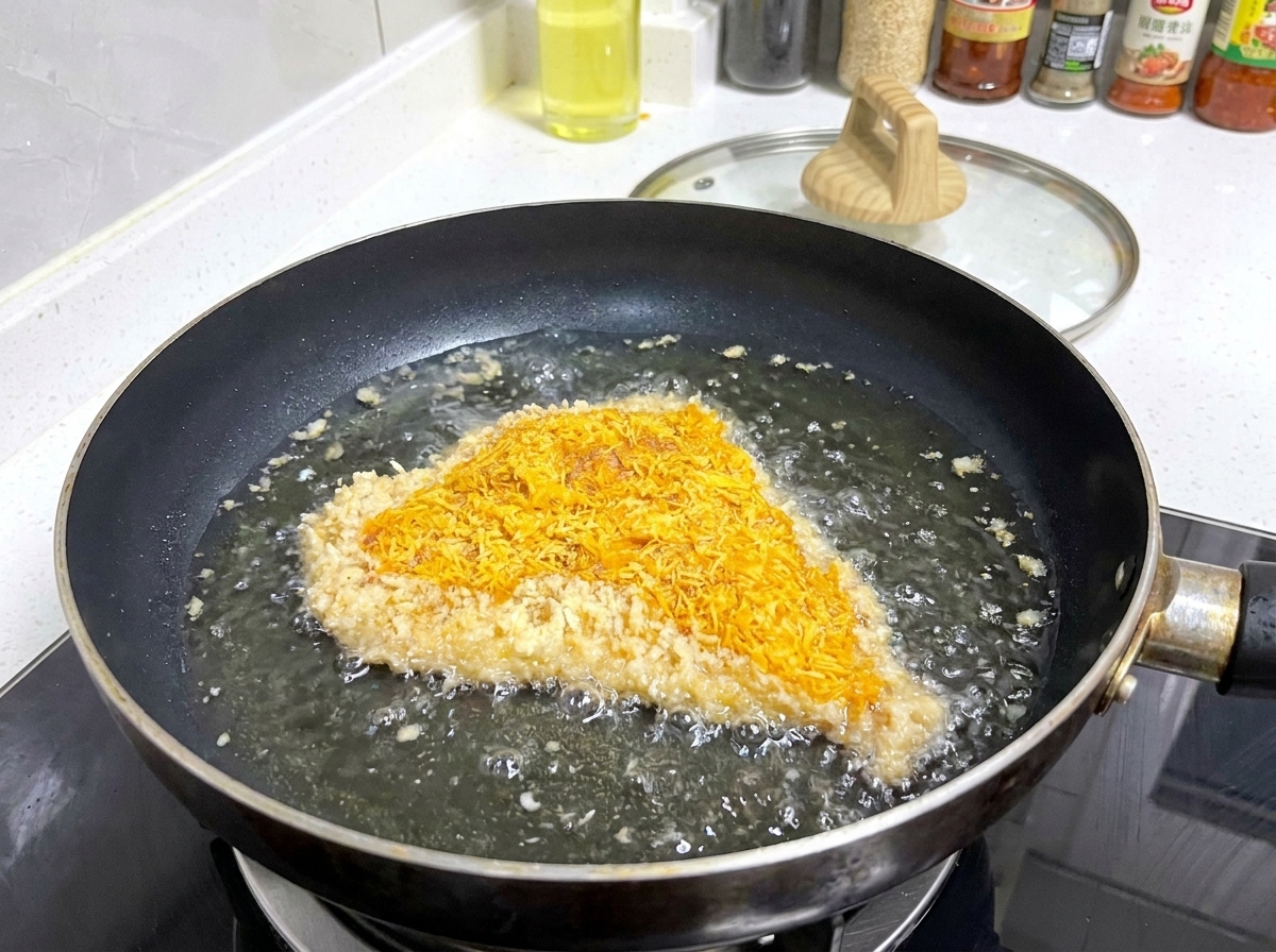 A breaded pork cutlet sizzling in a shallow pan of hot oil, with bubbles forming around the edges of the breading.