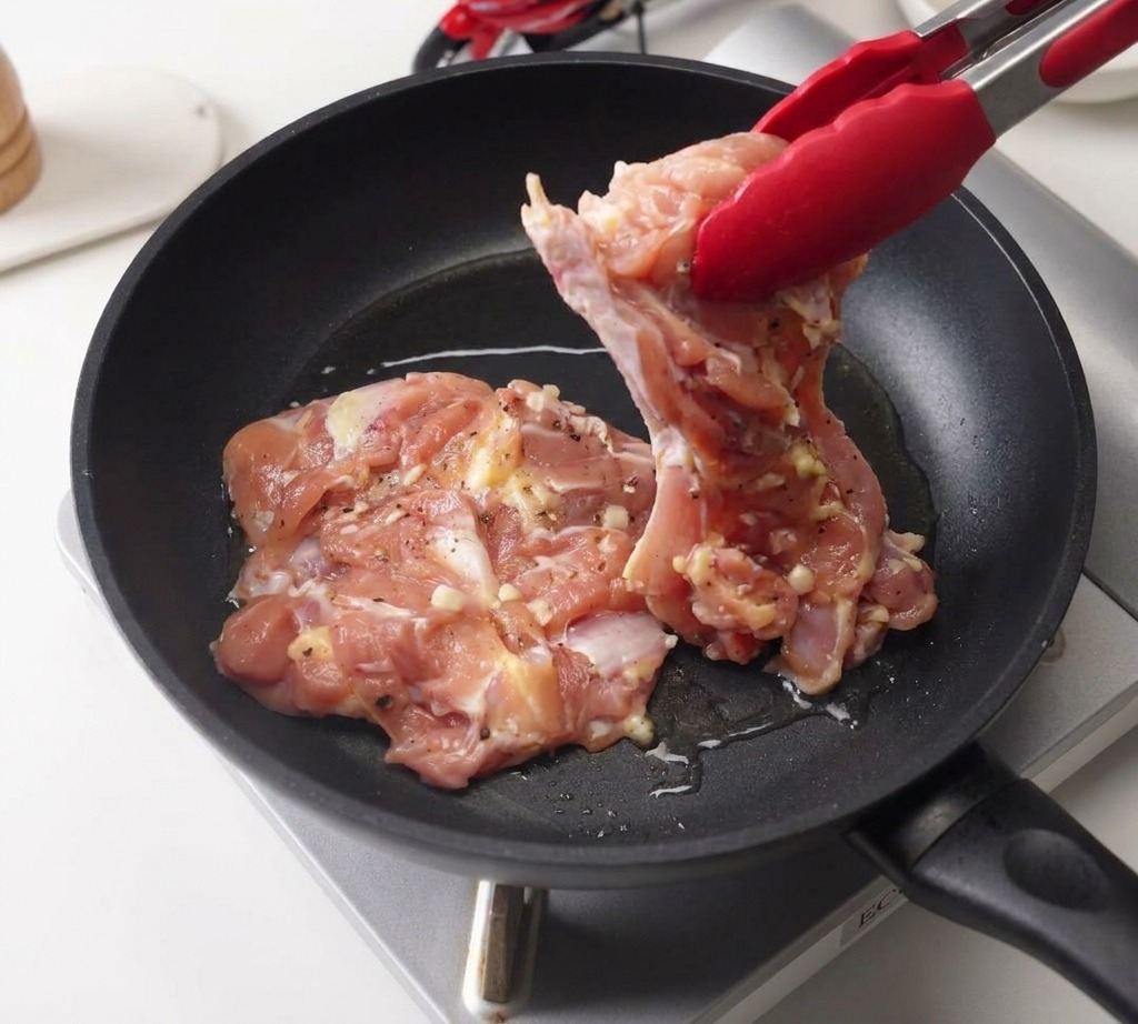 Red tongs placing a marinated raw chicken thigh into a black frying pan containing a thin layer of shimmering oil.