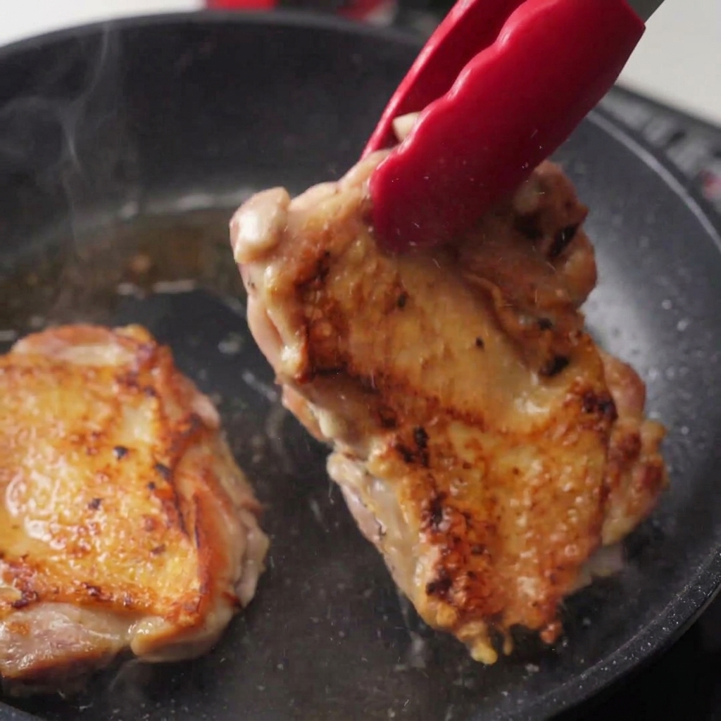 Red tongs lifting a fried chicken thigh in a pan, showing the underside which is deeply browned and crispy with visible steam rising.