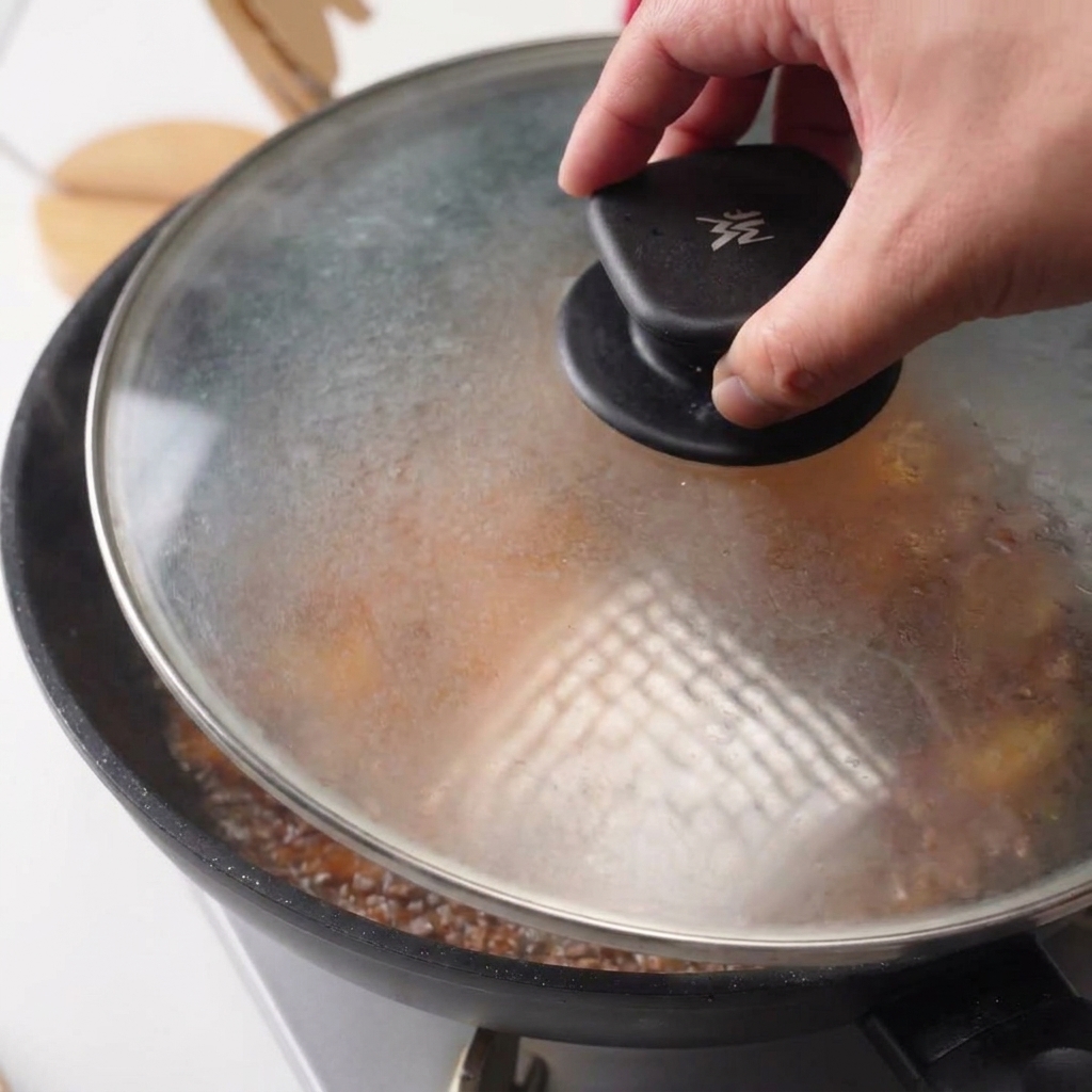 A hand placing a transparent glass lid onto a frying pan containing chicken and simmering sauce.