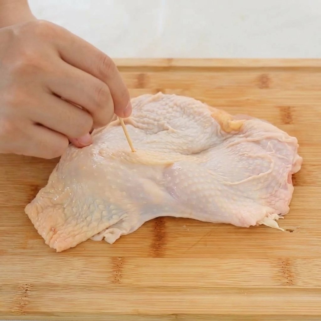 A close-up shot of a hand using a wooden toothpick to prick holes into the skin of a raw chicken thigh.
