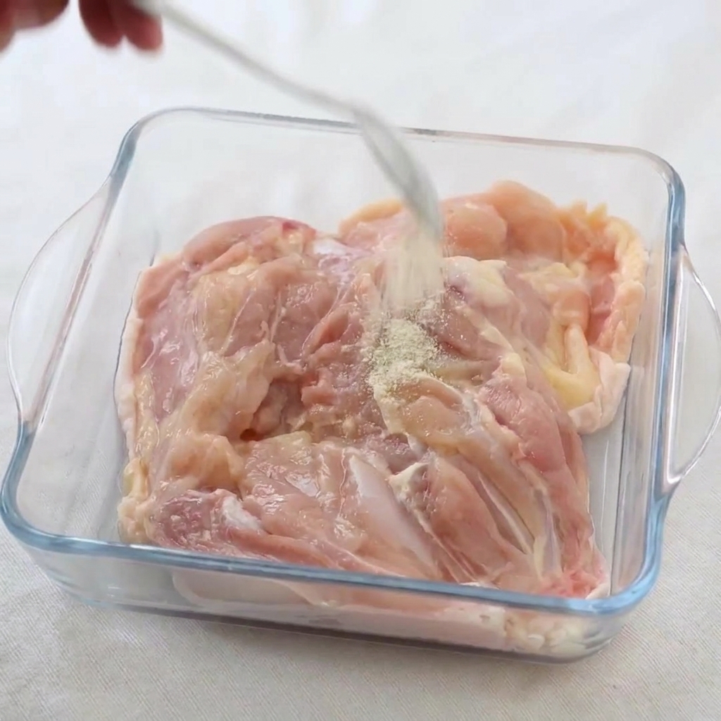 White pepper and salt being sprinkled from a spoon onto raw chicken thighs inside a glass dish.