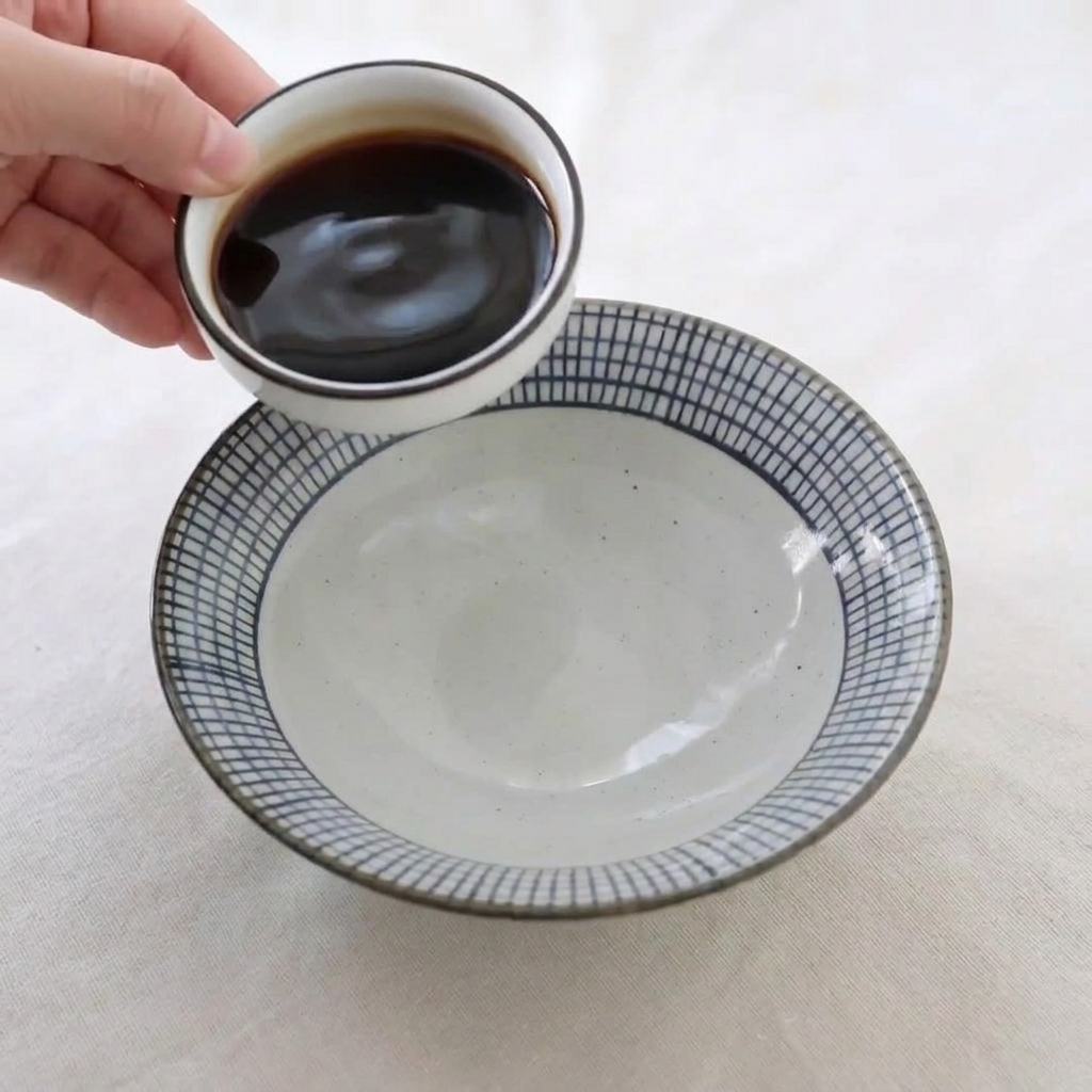 A hand pouring dark liquid seasonings from a small white dish into a larger ceramic bowl to prepare a honey glaze sauce.