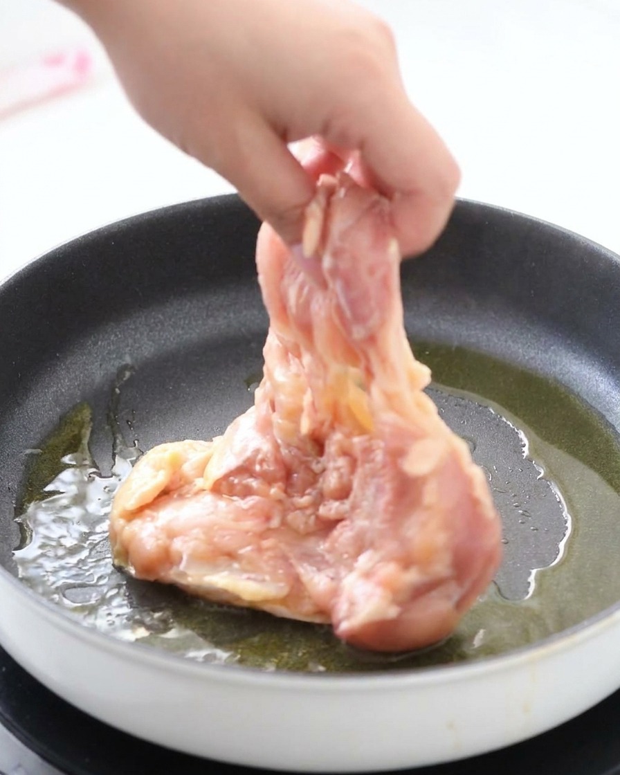 A hand placing a raw, marinated chicken thigh skin-side down into a hot frying pan with shimmering oil.