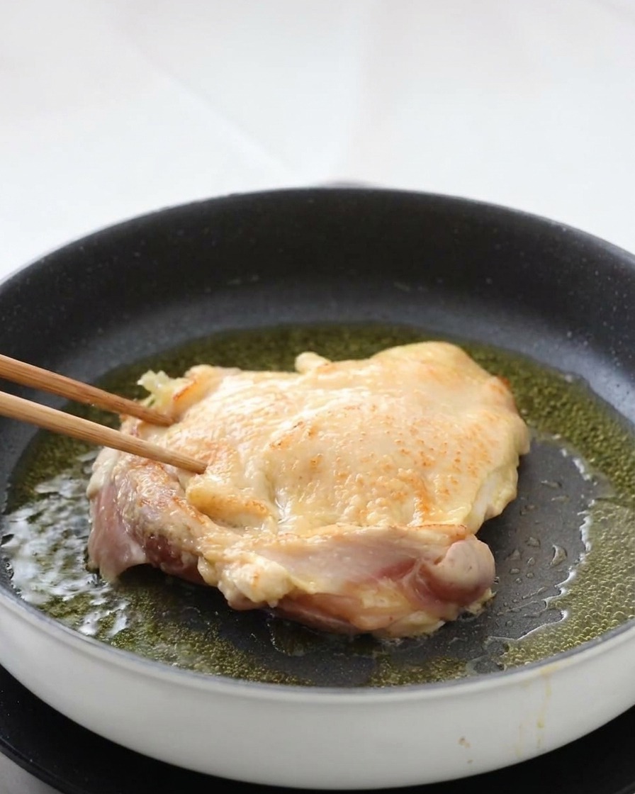 A chicken thigh being flipped in a pan with wooden chopsticks, revealing a perfectly browned and crispy skin surface.