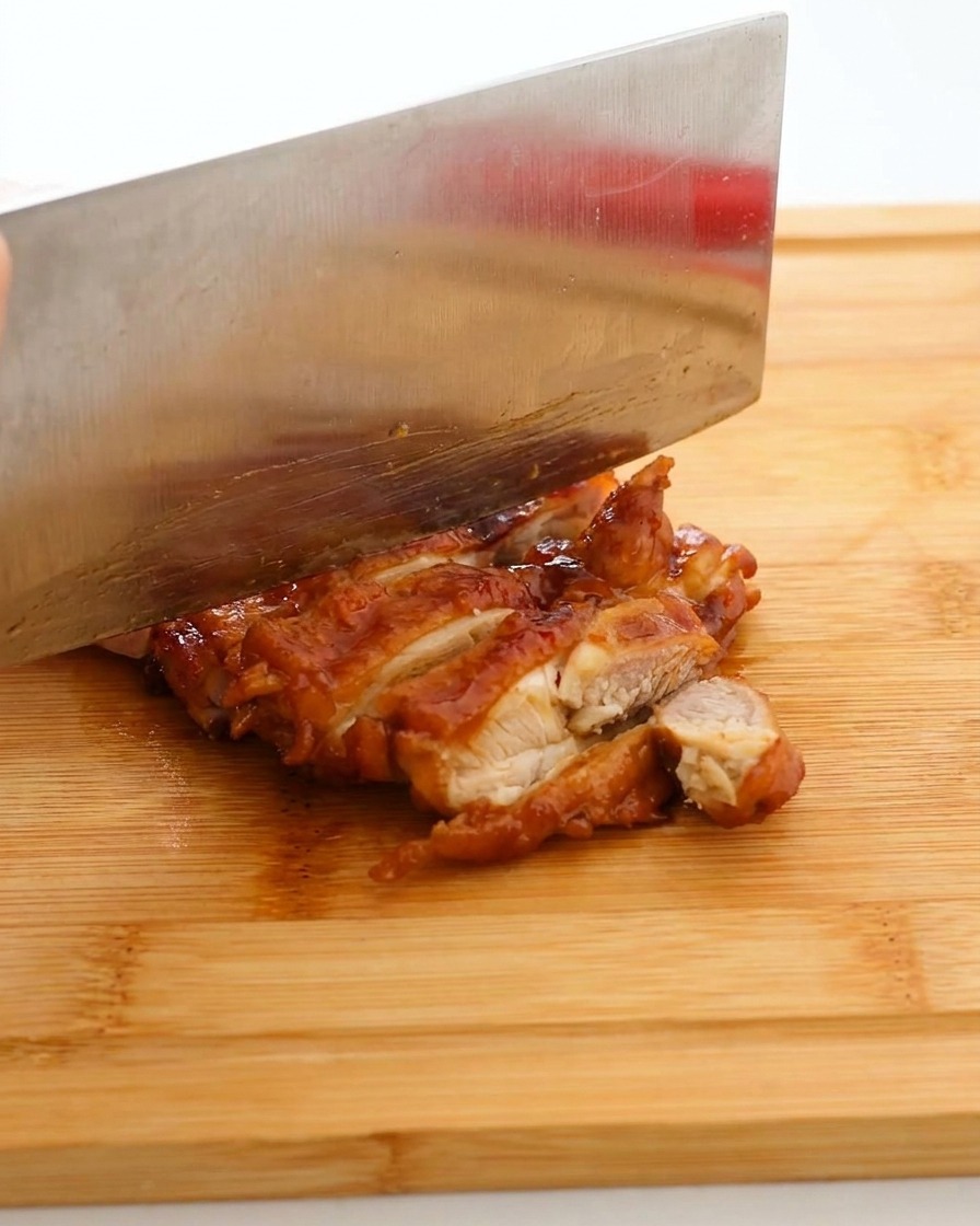 A large kitchen knife slicing a glistening, dark-glazed chicken thigh into strips on a wooden cutting board.