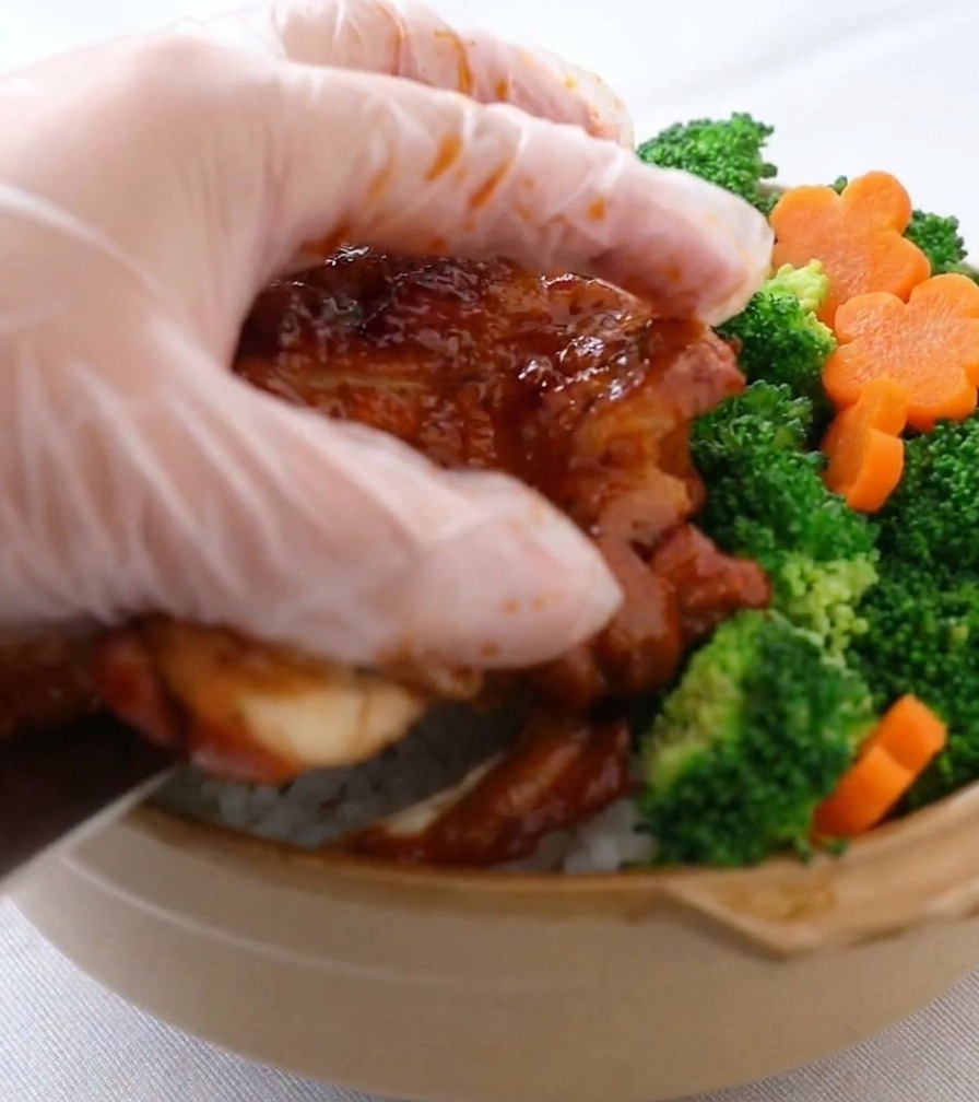 A hand wearing a clear plastic glove carefully placing bright green broccoli and orange carrot flowers into a ceramic bowl already filled with white rice and glazed chicken strips.