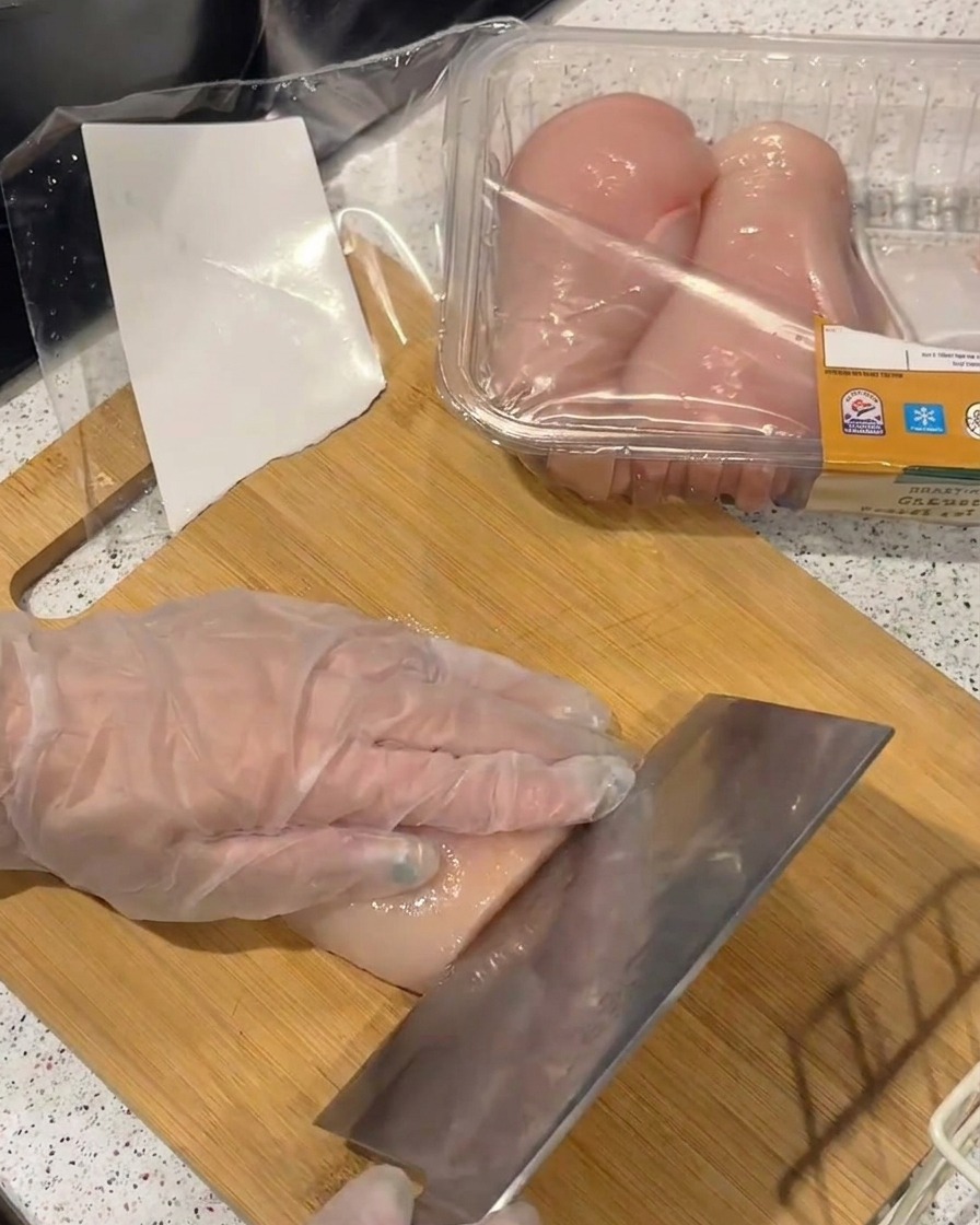 A hand in a clear plastic glove holding a raw chicken breast while slicing it into thin fillets with a large metal cleaver on a bamboo board.