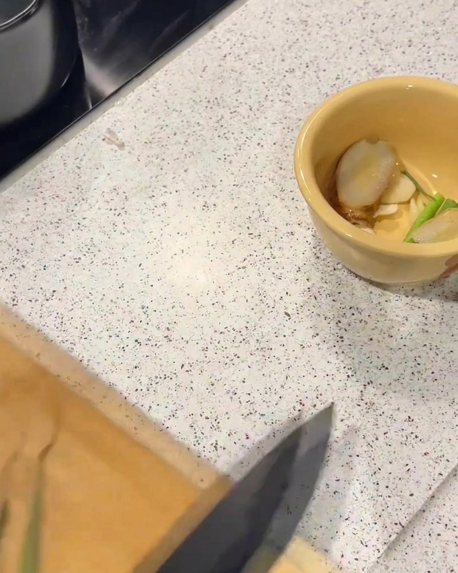A yellow ceramic bowl containing fresh ginger slices and green scallion pieces on a speckled countertop.