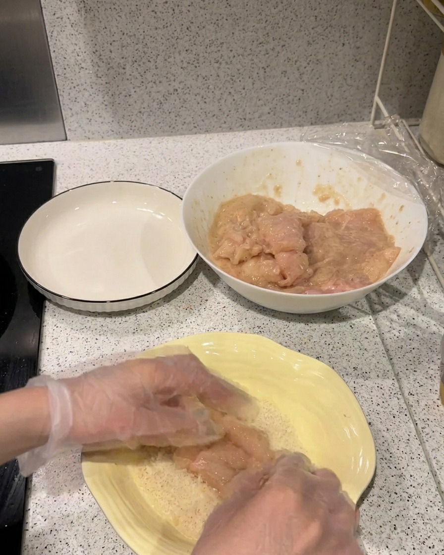 A marinated chicken fillet being pressed into a shallow yellow plate filled with light-colored Panko breadcrumbs.