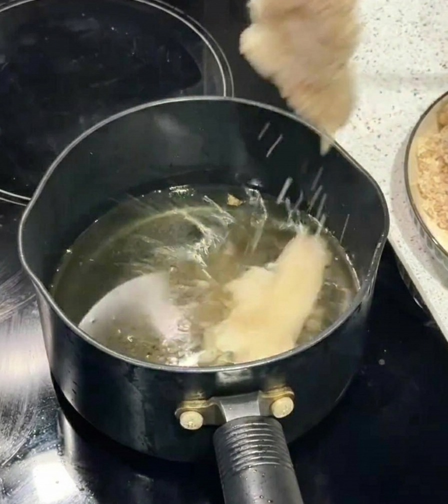 A hand using chopsticks to lower a breaded chicken fillet into a pot of bubbling cooking oil.