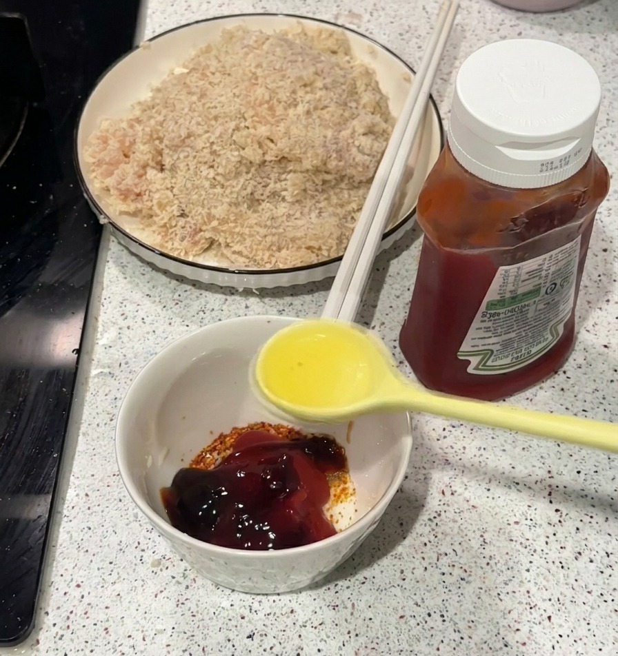 A yellow spoon pouring clear hot oil into a small white bowl containing ketchup and spices.