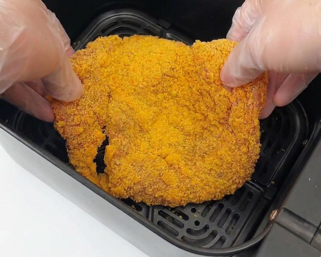 Two gloved hands placing a large piece of breaded chicken into the black basket of an air fryer.