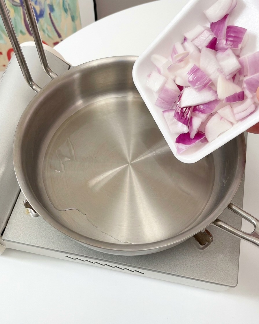 A hand pouring chopped red onions from a small square tray into a stainless steel pan with hot oil on a stove.