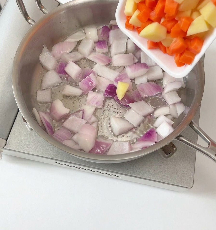 Diced potatoes and carrots being poured from a white tray into a stainless steel pan containing sautéed red onions.
